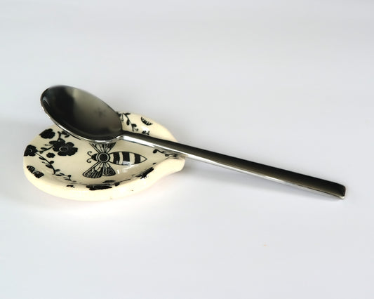 Round ceramic spoon plate with a bee design on a white surface and a spoon on top of it