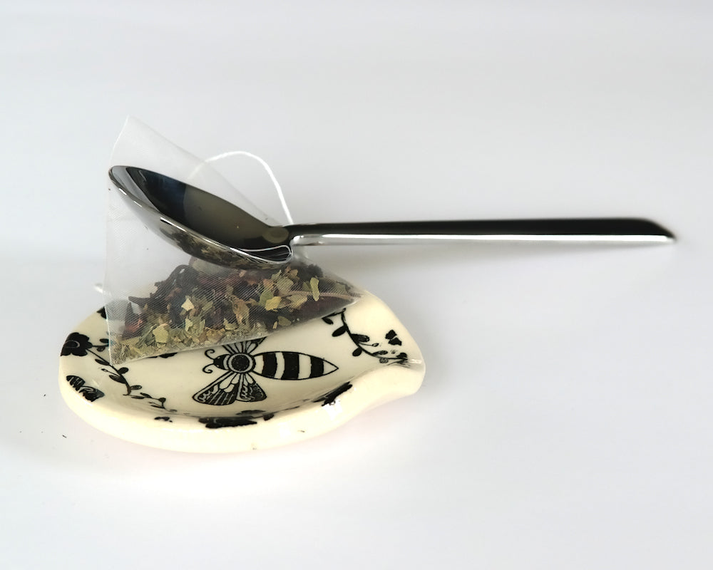 Round ceramic spoon plate with a bee design on a white surface and a spoon and a teabag on top of it