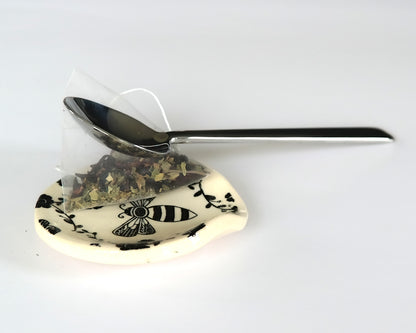 Round ceramic spoon plate with a bee design on a white surface and a spoon and a teabag on top of it