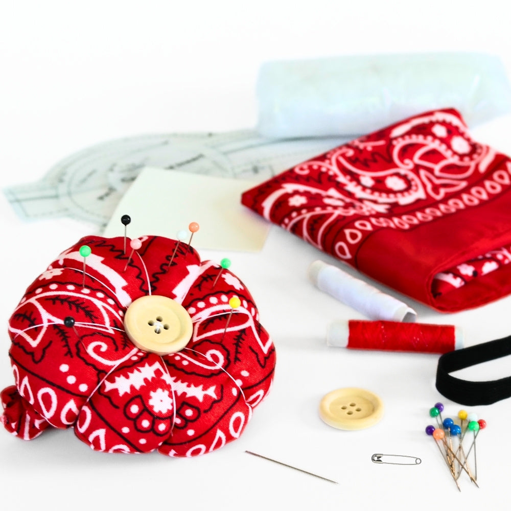 Finished red bandana flower pin cushion wristband with pins, shown with DIY sewing kit materials including thread, button, elastic, stuffing, and pattern pieces.