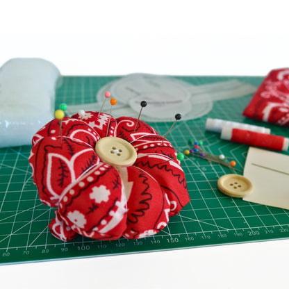 Finished red bandana flower pin cushion wristband with pins, shown with DIY sewing kit materials including thread, button, elastic, stuffing, and pattern pieces, with a lifestyle background. 