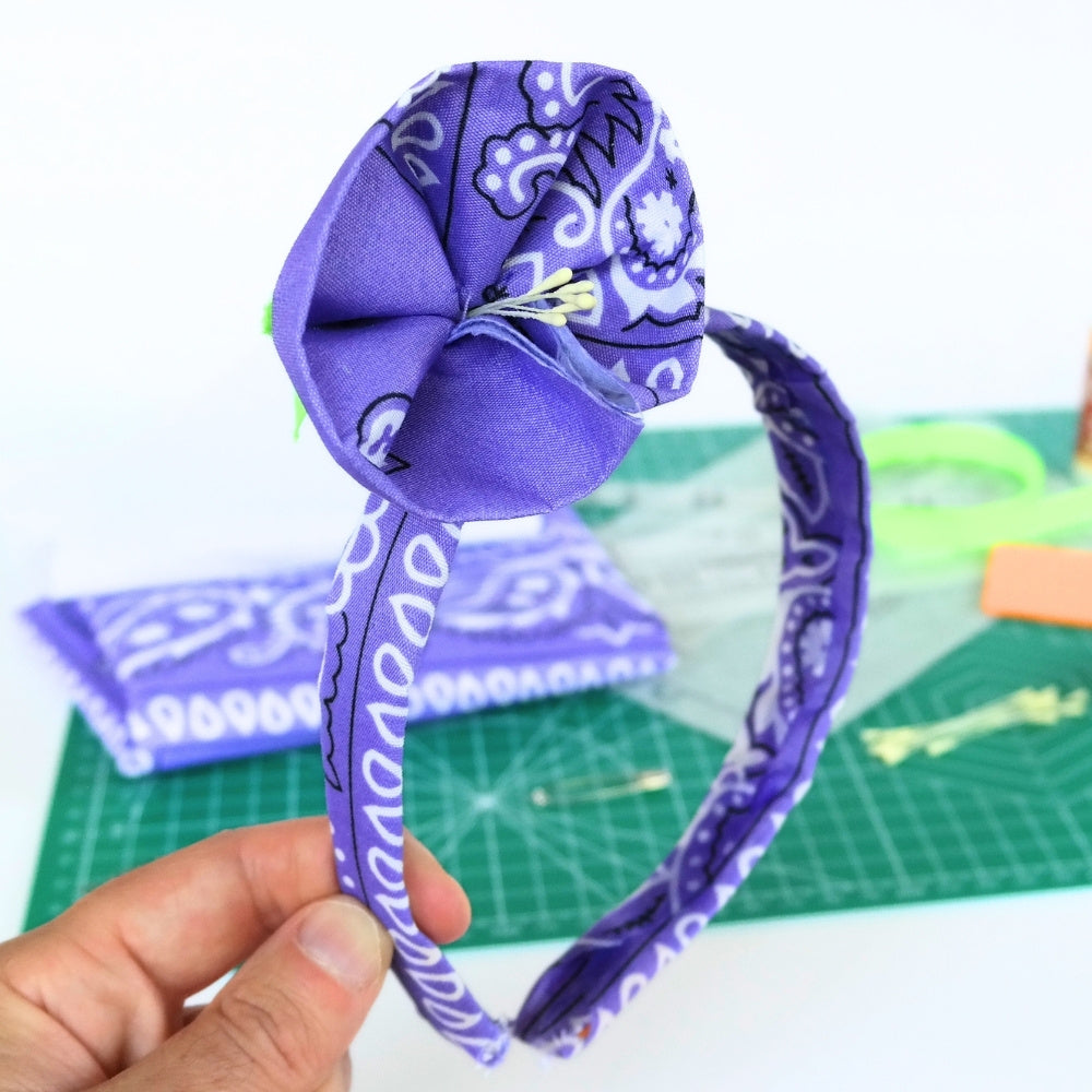Purple bandana headband with a flower detail attached in front of the DIY materials on a crafting mat used to make it.  