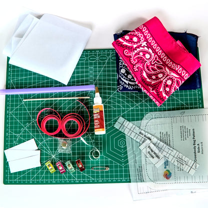 A flat-lay view of a quilting and fabric crafting kit on a white background, featuring a green cutting mat, white fabric pieces, a pink and navy paisley bandana, red fabric rolls, a purple stick, a bottle of fabric glue, ribbon, patterns, and other small crafting tools.
