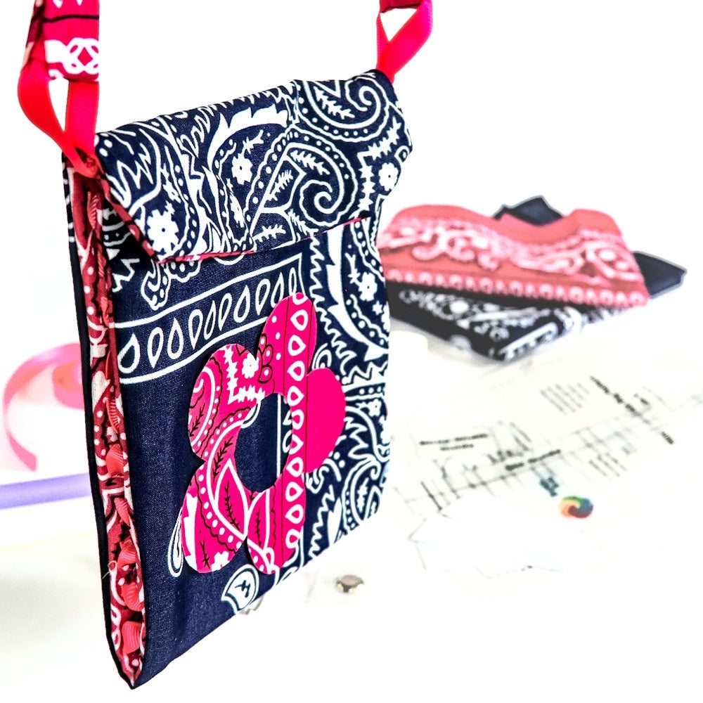A small fabric crossbody bag with a navy and white paisley print, a bright pink heart applique, and a red-and-white patterned strap, shown on a white background with crafting materials blurred in the background.