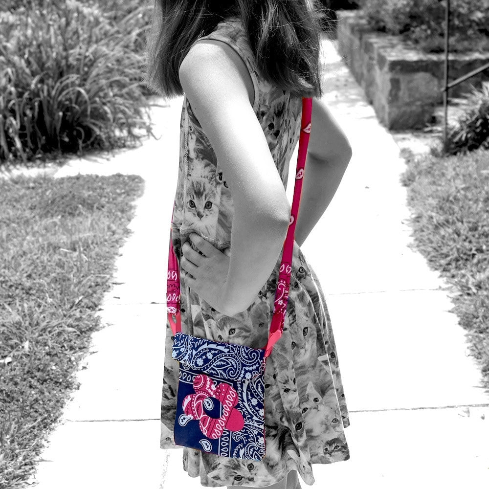 A girl standing on a sidewalk outdoors, wearing a sleeveless dress with a cat print and carrying a small crossbody bag with a blue, pink, and white bandana pattern. The photo is in black and white except for the bag and strap, which are in color.