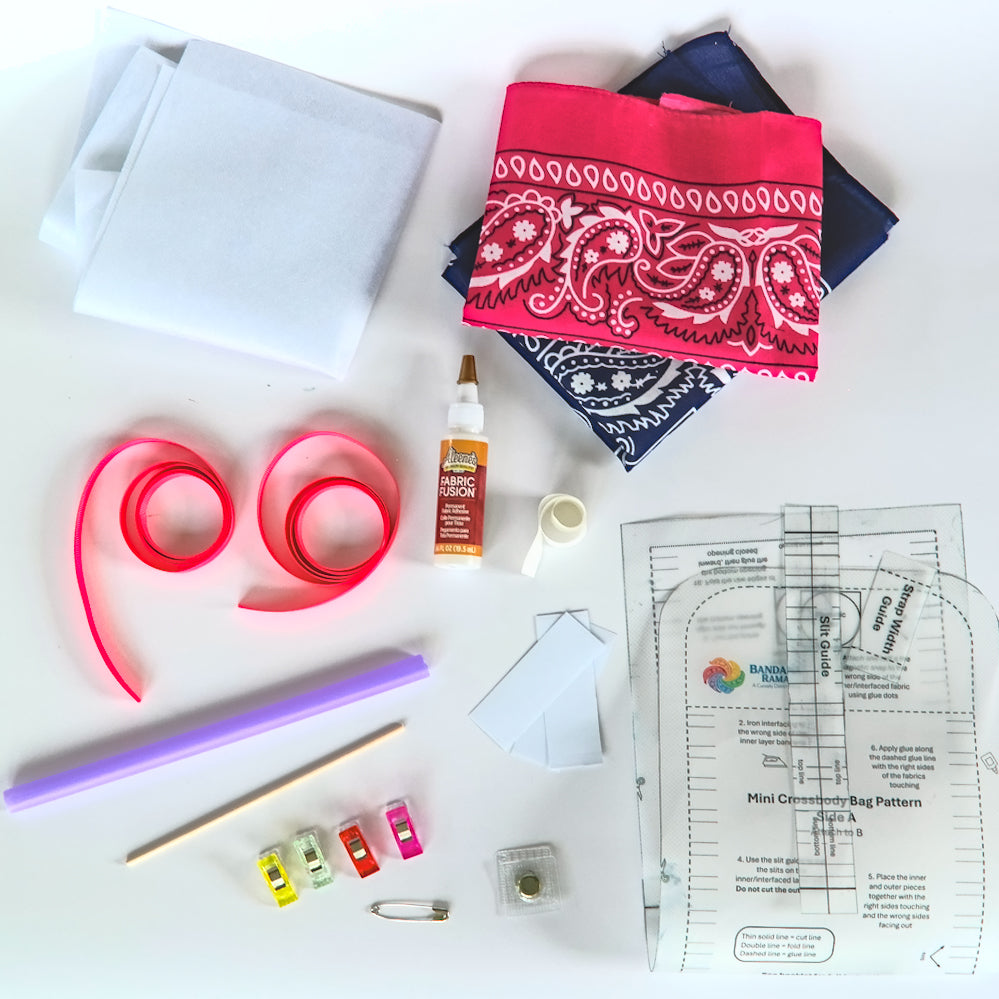 A flat-lay of sewing and crafting supplies on a white background, including folded white and patterned bandanas, pink ribbon, a bottle of fabric glue, a straw,  a wooden stick, small colored clips, a safety pin, and printed pattern templates.