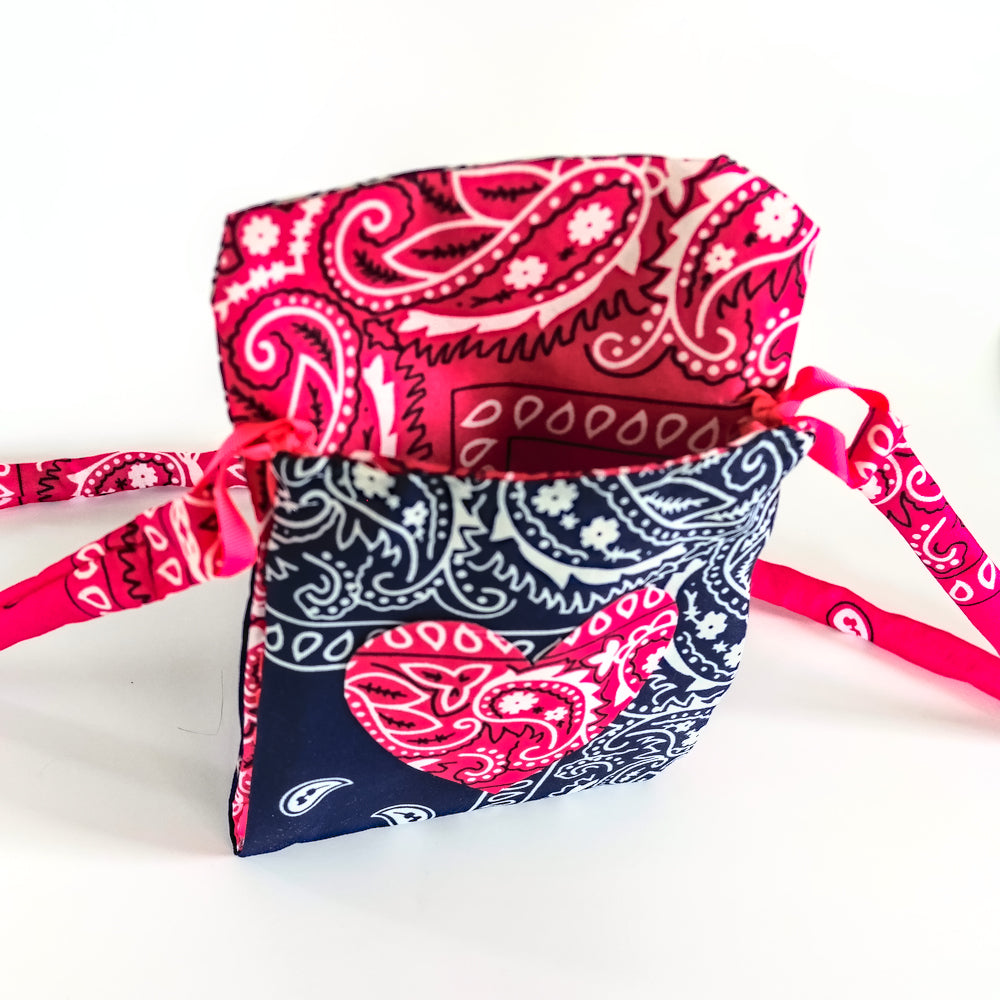 A small fabric bandana bag with a pink and navy blue paisley bandana print, featuring a top flap and adjustable tie straps, shown on a plain white background.
