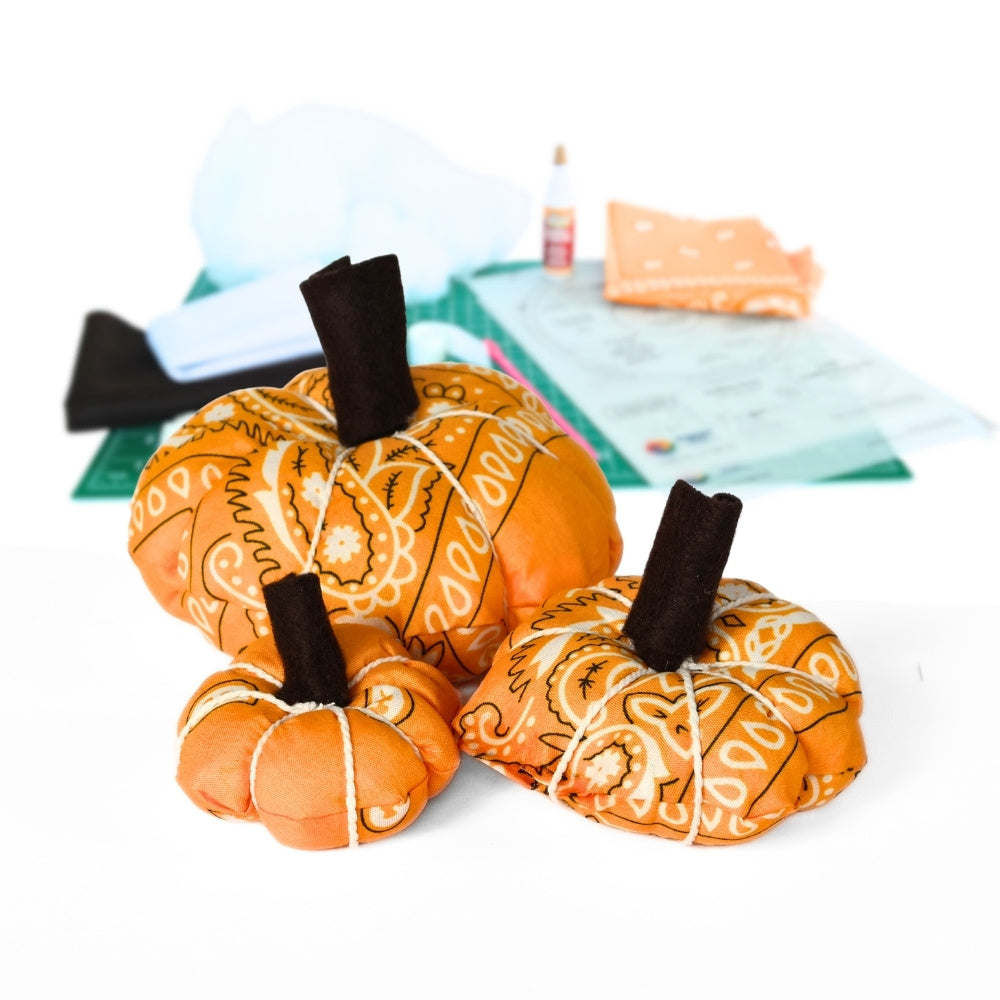 Three fabric pumpkins in orange paisley bandana patterns with dark brown stems, arranged on a white surface with sewing supplies in the background.